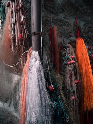 Several fishing nets in various colors including orange, red, and white are hanging against a rough stone wall. Some nets have floats attached, and ropes are tangled among them.