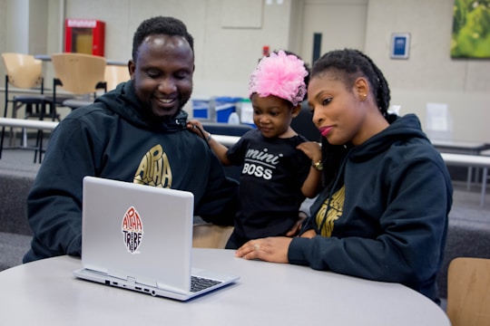 A family gathers around a laptop at a table in what appears to be a casual, open seating area. The man is smiling at the computer screen, while the woman and a small child, who is wearing a large pink flower headband and a shirt that says 'mini BOSS,' look on with interest. All three individuals are wearing dark hoodies with a logo on them.