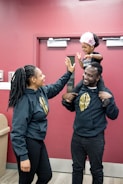 A joyful moment with a young child sitting on an adult's shoulders, reaching out for a high-five with another adult. All are wearing matching dark-colored hoodies with gold logos. The child is wearing a pink headband and smiling brightly. They stand in front of a maroon wall with a door.