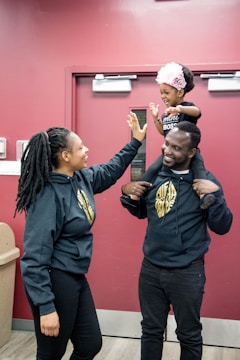 A joyful moment with a young child sitting on an adult's shoulders, reaching out for a high-five with another adult. All are wearing matching dark-colored hoodies with gold logos. The child is wearing a pink headband and smiling brightly. They stand in front of a maroon wall with a door.