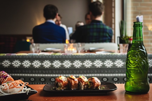 A plate of sushi is placed on a wooden table. The sushi looks freshly prepared, garnished with sauce and herbs. Next to the plate, there is an intricately designed green glass bottle with a depiction of a happy figure. In the background, people are seated at a dining establishment engaged in conversation. The atmosphere appears cozy with dim lighting and decorative patterns on the seating.