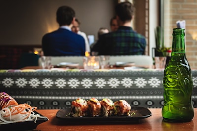 A plate of sushi is placed on a wooden table. The sushi looks freshly prepared, garnished with sauce and herbs. Next to the plate, there is an intricately designed green glass bottle with a depiction of a happy figure. In the background, people are seated at a dining establishment engaged in conversation. The atmosphere appears cozy with dim lighting and decorative patterns on the seating.