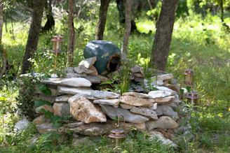 Кафяво‑сива каменна чешма, заобиколена от естествен каменен алпинеум в градина в Батак през деня