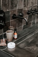 A bathroom countertop with black tiles featuring modern black wall-mounted faucets. Various toiletries are placed on the counter, including a gray cup, a glass, a brown wrapped package, a white wrapped object, and a bottle with a label. The overall setting is sleek and minimalistic.