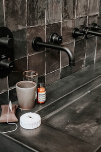 A bathroom countertop with black tiles featuring modern black wall-mounted faucets. Various toiletries are placed on the counter, including a gray cup, a glass, a brown wrapped package, a white wrapped object, and a bottle with a label. The overall setting is sleek and minimalistic.