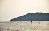 Two windsurfers move across a vast expanse of water, with a large, shadowy landmass forming the backdrop. The sky is overcast, casting a calm and muted light over the scene.