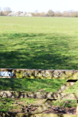 A wooden fence in a green field with the phrase 'BE KIND' and a heart symbol drawn in white chalk on one of the horizontal beams. The field stretches out under a clear blue sky, with trees lining the horizon in the background.
