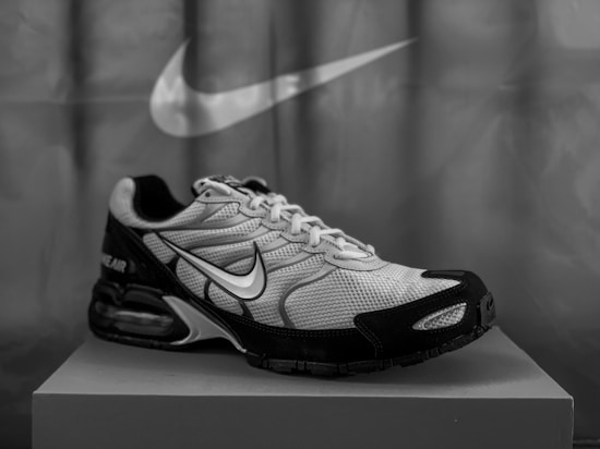 A black and white image of a single athletic sneaker positioned on a box, featuring a prominent logo on its side. The background displays a large, stylized brand logo.