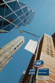 A cityscape viewed from below, featuring modern skyscrapers and a blue sky. Geometric glass architecture is prominent, with a streetlight and a bus stop sign in the foreground, indicating routes M20 and M104, and locations Lincoln Center and West Harlem.
