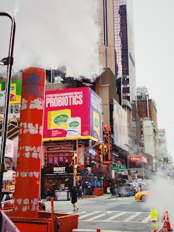A bustling urban street scene featuring towering buildings with colorful billboards, including a prominent advertisement for probiotics. Steam rises from a street vent near an orange barricade. Cars and pedestrians, including a woman in black, are visible navigating the busy intersection.