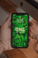 A hand holding a smartphone displaying a lock screen with a background of green ferns and the words 'ONE Goal. Purpose'. The time shown is 5:32 on Wednesday, 25 March. An Apple computer is visible in the background.