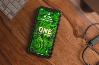 A smartphone with a green fern-themed wallpaper displaying the words 'ONE GOAL, PURPOSE' rests on a wooden table. The time on the phone reads 5:25, with the date being Wednesday, 25 March. Nearby are part of a keyboard and a coiled cable.