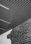 Combination of a corrugated metal ceiling and a part brick, part plastered wall. The brick wall has an unfinished, rough edge where the plaster meets the brick. The ceiling features linear fluorescent light fixtures.
