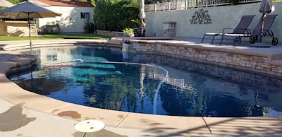 A serene outdoor swimming pool surrounded by a stone patio and lounge chairs. The water is clear and reflects the surrounding greenery and structures. An umbrella provides shade nearby, and there's a privacy fence with greenery in the background. The area is well-maintained and sunlit, suggesting a peaceful and relaxing setting.