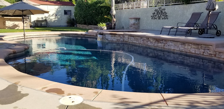 A serene outdoor swimming pool surrounded by a stone patio and lounge chairs. The water is clear and reflects the surrounding greenery and structures. An umbrella provides shade nearby, and there's a privacy fence with greenery in the background. The area is well-maintained and sunlit, suggesting a peaceful and relaxing setting.