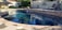 A serene outdoor swimming pool surrounded by a stone patio and lounge chairs. The water is clear and reflects the surrounding greenery and structures. An umbrella provides shade nearby, and there's a privacy fence with greenery in the background. The area is well-maintained and sunlit, suggesting a peaceful and relaxing setting.