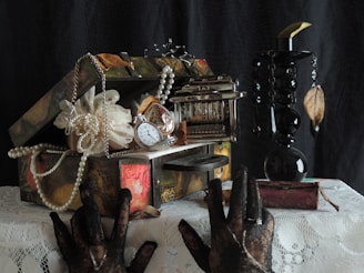 An ornate, vintage box overflowing with pearl necklaces, a pocket watch, and decorative items sits on a lace tablecloth. Adjacent is a tall, dark, ornamental vase with dangling beads and dried leaves. Two hands in lace gloves reach towards the objects.