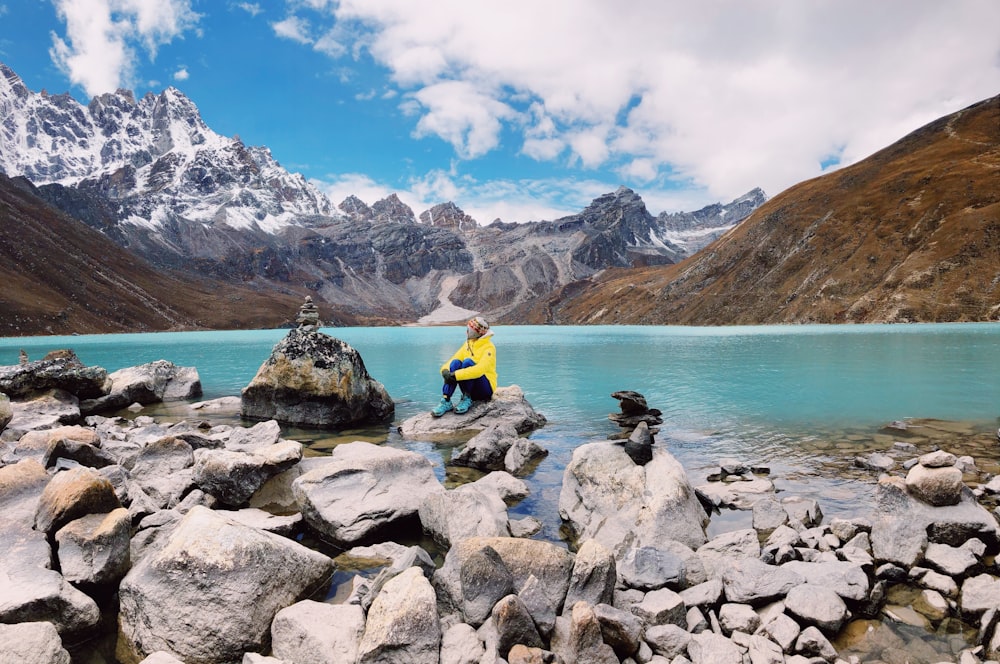 man in yellow shirt sitting on rock near body of water during daytime