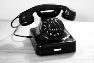 A vintage rotary dial telephone is placed on a wooden surface. The phone is black with a circular dial featuring numbers and letters. Its handset rests on top of the main body, connected by a coiled cord.