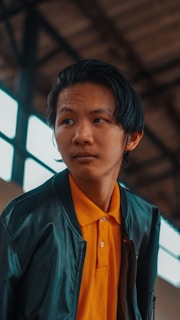 A young person with dark hair wearing a black jacket over a bright orange collared shirt. The background is an industrial setting with blurred metal beams and windows.