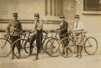 Four young boys are standing with their bicycles in front of a stone building. They are wearing formal attire with caps and long socks. The building features large columns and intricate architectural details.