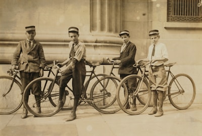 Four young boys are standing with their bicycles in front of a stone building. They are wearing formal attire with caps and long socks. The building features large columns and intricate architectural details.