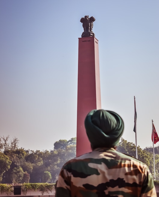 A person wearing a camouflage uniform and a green turban stands in front of a tall red monument topped with a sculpture. Flags are visible in the background, along with green trees under a clear blue sky.