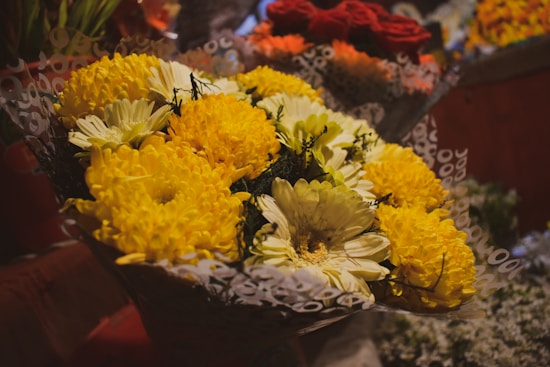 A bouquet of vibrant yellow and white flowers wrapped in decorative paper, with a background featuring additional floral arrangements in various colors.