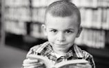 A young child with a neutral expression is holding a book while looking directly at the camera. The background features a blurred bookshelf filled with books, suggesting a library setting. The child is wearing a checkered shirt, and the photograph is in black and white.
