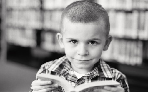A young child with a neutral expression is holding a book while looking directly at the camera. The background features a blurred bookshelf filled with books, suggesting a library setting. The child is wearing a checkered shirt, and the photograph is in black and white.