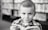 A young child with a neutral expression is holding a book while looking directly at the camera. The background features a blurred bookshelf filled with books, suggesting a library setting. The child is wearing a checkered shirt, and the photograph is in black and white.