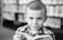 A young child with a neutral expression is holding a book while looking directly at the camera. The background features a blurred bookshelf filled with books, suggesting a library setting. The child is wearing a checkered shirt, and the photograph is in black and white.