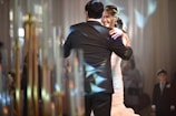 A couple is dancing together in a formal setting, where the woman is wearing a white dress and the man is in a black suit. The scene is warmly lit with reflections of light creating patterns on their attire. Onlookers, including a child in a suit, are blurred in the background amidst soft-focus lighting and elegant decor.