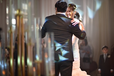 A couple is dancing together in a formal setting, where the woman is wearing a white dress and the man is in a black suit. The scene is warmly lit with reflections of light creating patterns on their attire. Onlookers, including a child in a suit, are blurred in the background amidst soft-focus lighting and elegant decor.
