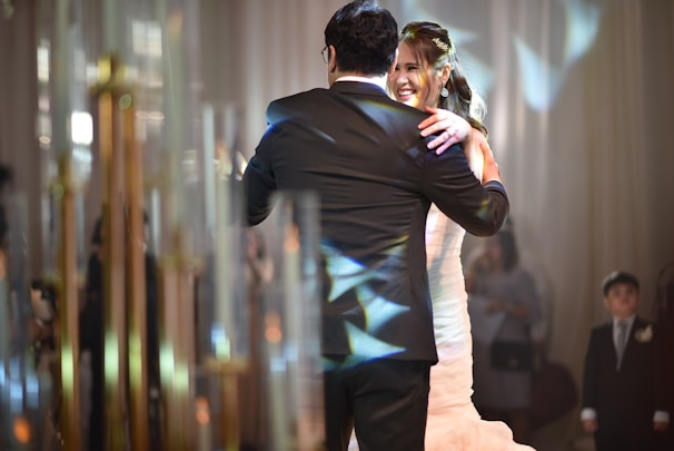 A couple is dancing together in a formal setting, where the woman is wearing a white dress and the man is in a black suit. The scene is warmly lit with reflections of light creating patterns on their attire. Onlookers, including a child in a suit, are blurred in the background amidst soft-focus lighting and elegant decor.