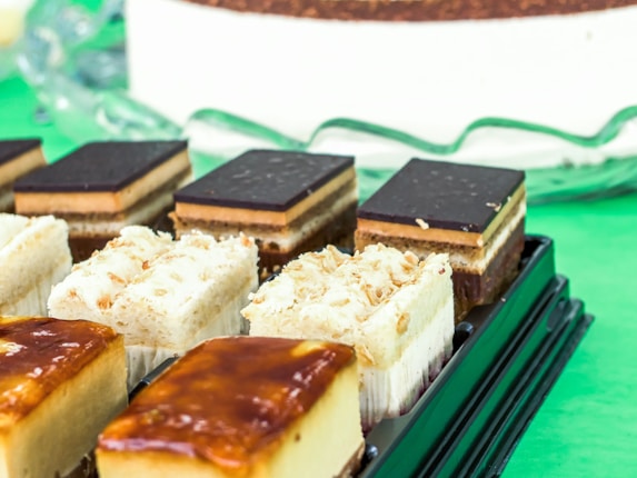 An assortment of layered desserts is displayed on a black tray. The desserts have various layers including creamy fillings and dark chocolate toppings. The background features a large circular dessert and a bright green surface.