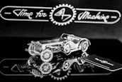 A highly detailed, metallic model of a vintage car is positioned on a reflective surface. The intricate design showcases gears and a classic car body. The background features the phrase 'Time for Machine' in a stylized script, offering a sense of branding or theme.