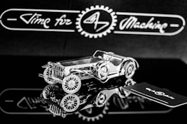 A highly detailed, metallic model of a vintage car is positioned on a reflective surface. The intricate design showcases gears and a classic car body. The background features the phrase 'Time for Machine' in a stylized script, offering a sense of branding or theme.
