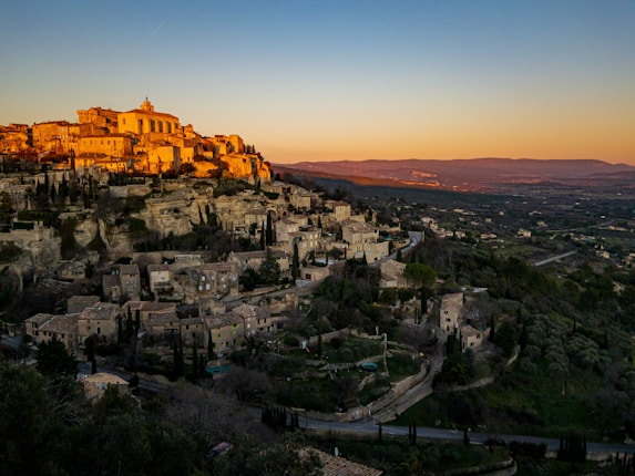 A scenic view of a picturesque village built on a hillside, basking in the warm hues of a setting sun. The village architecture features stone buildings with red tile roofs, surrounded by lush greenery and cypress trees. The expansive landscape in the background stretches towards distant mountains under a gradient sky transitioning from warm tones to cooler blues.