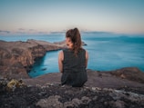 woman in black tank top sitting on rock near body of water during daytime