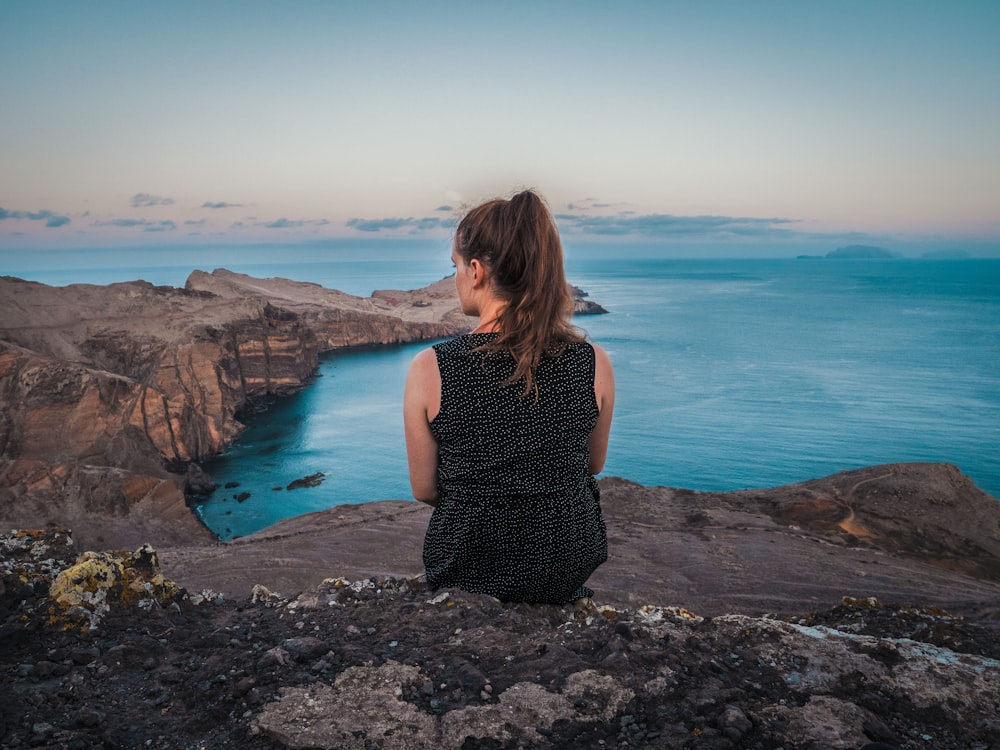 woman in black tank top sitting on rock near body of water during daytime