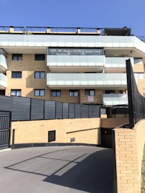 A modern multi-story residential building with light brick walls and balconies with translucent glass railings. There are signs or banners displayed on one of the balconies. The foreground features a paved driveway leading to an underground parking area. The sky is clear and blue.