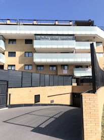 A modern multi-story residential building with light brick walls and balconies with translucent glass railings. There are signs or banners displayed on one of the balconies. The foreground features a paved driveway leading to an underground parking area. The sky is clear and blue.