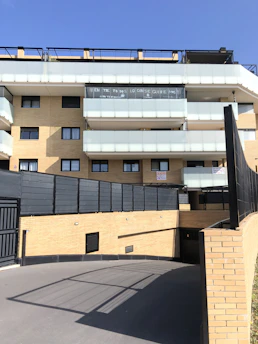 A modern multi-story residential building with light brick walls and balconies with translucent glass railings. There are signs or banners displayed on one of the balconies. The foreground features a paved driveway leading to an underground parking area. The sky is clear and blue.