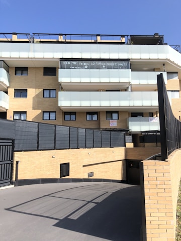 A modern multi-story residential building with light brick walls and balconies with translucent glass railings. There are signs or banners displayed on one of the balconies. The foreground features a paved driveway leading to an underground parking area. The sky is clear and blue.