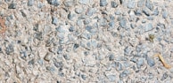 A close-up view of a rough concrete surface embedded with small, irregularly shaped stones, predominantly gray in color. The texture is uneven, showcasing a mix of pebbles and cement.