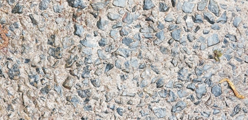 A close-up view of a rough concrete surface embedded with small, irregularly shaped stones, predominantly gray in color. The texture is uneven, showcasing a mix of pebbles and cement.
