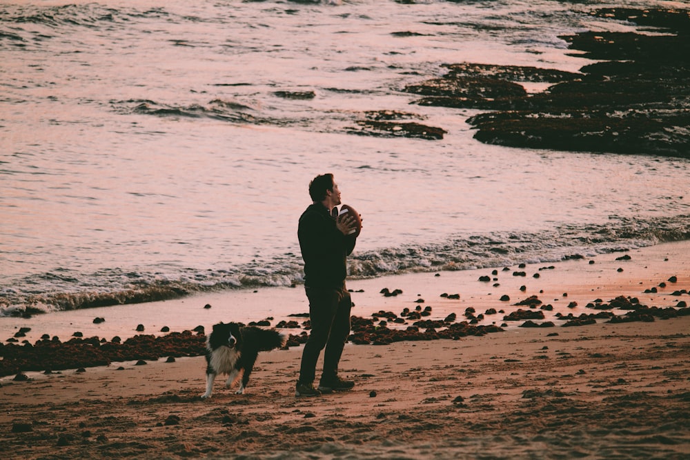 man in black jacket standing on seashore with dog