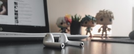 A pair of white wireless earbuds lie on a dark surface. In the background, a laptop screen displays some text, while three toy figures and a small plant in a pot are slightly out of focus.