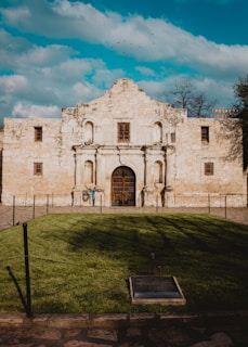 A historic stone building with an ornate facade and arched doorway. The architecture features several small, square windows and aged textures. It is set against a bright blue sky with some fluffy clouds and a small flock of birds. In the foreground, well-maintained green grass is framed by a low rope barrier, and a plaque is embedded in the ground.